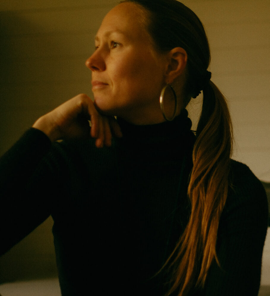 A woman with long light brown hair in a ponytail, wearing a black turtleneck and large hoop earrings, looking thoughtfully to the side in a dimly lit room with a sewing machine in the background.